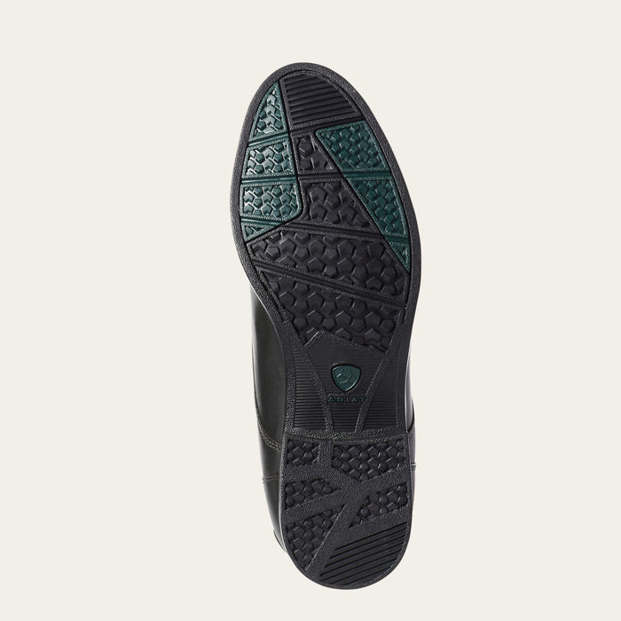 The outsole of a black leather shoe, showing textured tread patterns and green grip accents, photographed from a top-down view on a light beige background.
