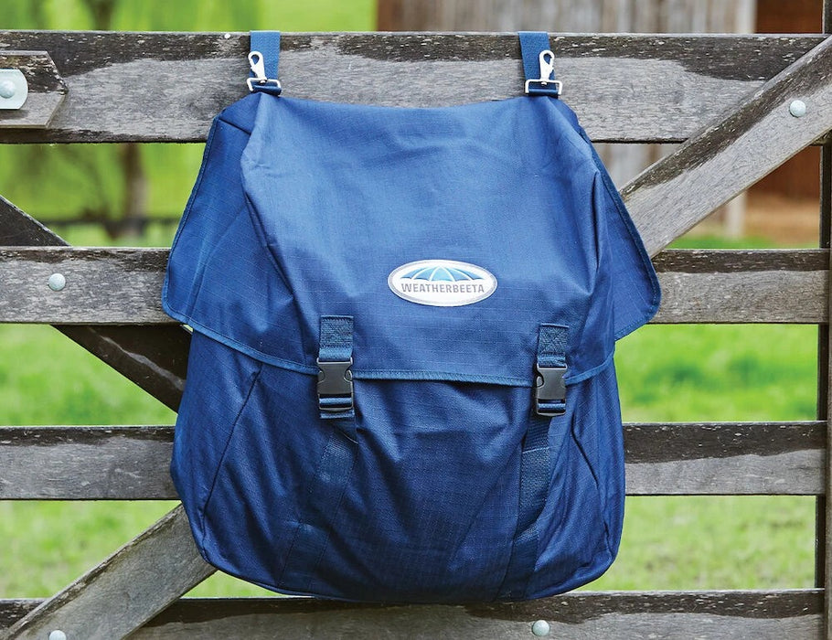 A blue waterproof blanket bag hanging on a weathered wooden fence outdoors, with a visible WeatherBeeta logo patch on the front flap.