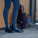 A child wearing dark navy equestrian tights and shiny black Dublin paddock riding boots stands beside a brown leather saddle in front of a wooden stable wall.