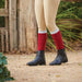 A person outdoors wearing beige riding tights, bright red knee-high socks, and black ankle paddock boots on a dirt path with a rustic wooden fence and greenery in the background.