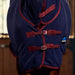 A close-up of the front of a horse wearing a navy blue cooler blanket and hood with burgundy trim and straps, photographed in a stable setting.