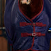 A close-up of a horse wearing a navy blue cooler with burgundy trim and double buckle straps, set against a dark stable background.