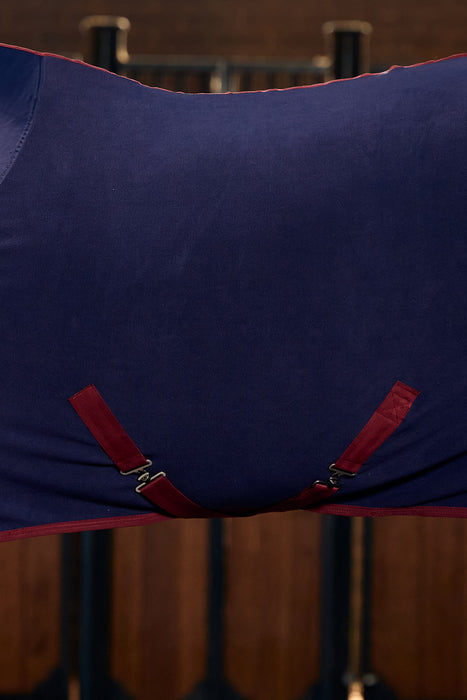 A close up of a navy cooler with burgundy surcingles crossed under the belly of a horse that is standing in a barm