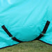 A close-up of a turquoise blue horse blanket with black surcingle straps crossed under the belly and trim, shown outdoors on a grassy lawn with a wooden fence in the background.