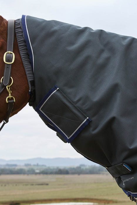 A close-up side view of a horse wearing a dark gray turnout blanket with navy and white trim, along with a dark bridle, set against an outdoor landscape background.