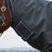 A close-up side view of a horse wearing a dark gray turnout blanket with navy and white trim, along with a dark bridle, set against an outdoor landscape background.