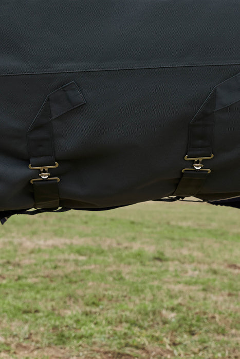 Close-up of a black waterproof horse blanket with crossing sircingles and brass buckle straps, shown outdoors over a grassy field.