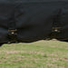 Close-up of a black waterproof horse blanket with crossing sircingles and brass buckle straps, shown outdoors over a grassy field.