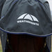 A close-up of a horse turnout blanket with an attached neck cover, shown on a horse from behind. The cover is dark gray with a white Weatherbeeta logo and white piping, with blue trim along the edge.