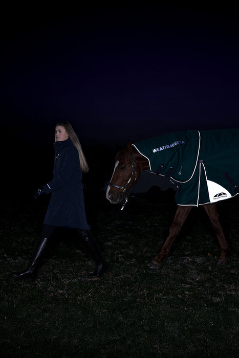A woman in a dark navy coat walks beside a horse wearing a dark green Weatherbeast turnout blanket with white trim and logo, set against a pitch-black night background. Showing off the reflective coating on the blanket 