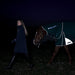 A woman in a dark navy coat walks beside a horse wearing a dark green Weatherbeast turnout blanket with white trim and logo, set against a pitch-black night background. Showing off the reflective coating on the blanket 