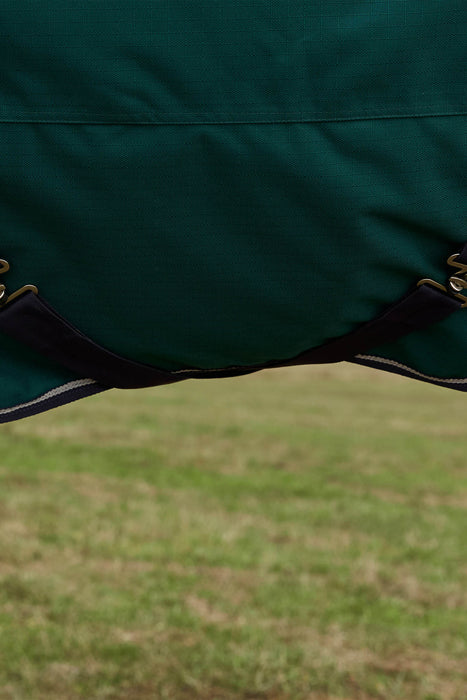 Close-up of a dark green fabric horse blanket with black surcingles crossing under the horses belly and silver tone metal hardware, shown outdoors over a blurred grassy field.