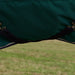Close-up of a dark green fabric horse blanket with black surcingles crossing under the horses belly and silver tone metal hardware, shown outdoors over a blurred grassy field.