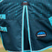 Close-up of a horse blanket with a dark teal patterned upper section, bright turquoise piping, and a Weatherbeta logo patch on the lower right.