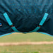 Close-up of a dark teal patterned fabric turnout blanket with bright teal surcingles and trim, photographed outdoors against a blurred grassy field.