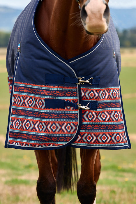 A close-up of a horse wearing a navy blue turnout blanket with a red, white, and blue geometric patterned lower panel, set against a blurred outdoor field background.