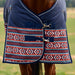 A close-up of a horse wearing a navy blue turnout blanket with a red, white, and blue geometric patterned lower panel, set against a blurred outdoor field background.