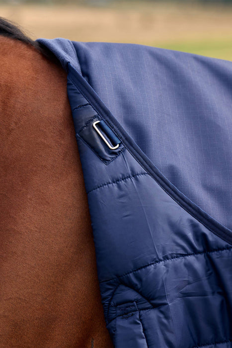Close-up of a navy blue quilted horse blanket or tack garment with a silver metal D-ring attachment, shown on a brown horse against an outdoor blurred background.