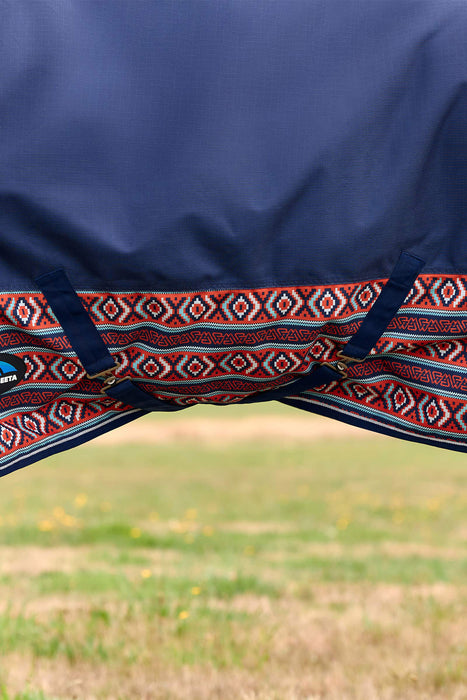 Close-up of a navy blue turnout blanket for a horse with a red, white, and blue geometric patterned hem with 2 surcingles attached under the belly, shown outdoors with a blurred grassy field in the background.