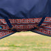 Close-up of a navy blue turnout blanket for a horse with a red, white, and blue geometric patterned hem with 2 surcingles attached under the belly, shown outdoors with a blurred grassy field in the background.