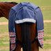 A navy blue horse turnout blanket with red, white, and blue geometric patterned trim, shown on the back of a brown horse in an outdoor grassy field.