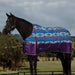 A dark brown horse standing in a grassy field under a cloudy sky, wearing a patterned horse blanket with blue, white, and purple Southwestern-style geometric designs and purple lower panels.