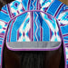 A close-up of a horse's rump and tail area wearing a patterned horse blanket or tack cover. The fabric has a geometric Southwestern-style print in blue, purple, and white, with a gray reflective strip along the front edge.