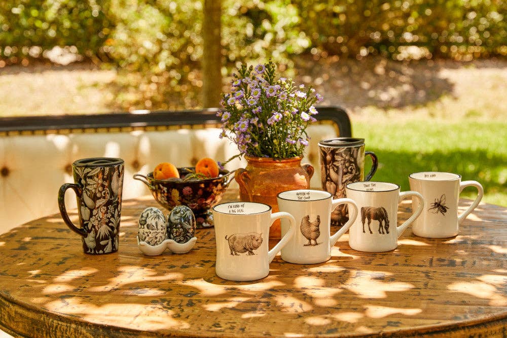 A collection of ceramic mugs with rustic animal illustrations, displayed on a wooden table outdoors with a blurred garden background.