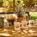 A collection of ceramic mugs with rustic animal illustrations, displayed on a wooden table outdoors with a blurred garden background.