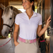 A woman in a white polo shirt and beige riding pants stands in a stable next to a gray horse, wearing a pink belt with a silver horseshoe buckle.