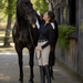 A woman in equestrian attire, including a black long-sleeve shirt, beige riding pants, and black knee-high boots, stands beside a black horse in an outdoor stable setting.