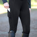 Black equestrian riding breeches with a side pocket holding a white phone, worn with a black leather belt and tall black riding boots on an outdoor path.