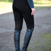 A person outdoors wearing black riding breeches with a subtle textured panel near the lower leg, paired with a dark long-sleeve top and tall black riding boots.