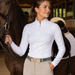 A woman wearing a white long-sleeve quarter-zip equestrian shirt and beige riding pants stands beside a brown horse in a stable, holding the horse's reins.