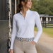 A woman wearing a white long-sleeve quarter-zip performance shirt and beige riding pants, standing outdoors near a stable with a patterned belt.