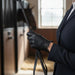 A woman in a navy equestrian jacket and black riding glove holds a black riding crop with a leather strap inside an indoor stable corridor.