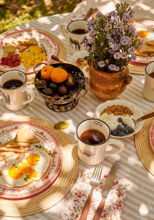 A styled breakfast table outdoors with plates of eggs, toast, fruit, coffee, and a small vase of purple flowers on a striped tablecloth.