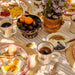 A styled breakfast table outdoors with plates of eggs, toast, fruit, coffee, and a small vase of purple flowers on a striped tablecloth.