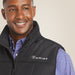 A smiling man wearing a black Ariat zip-front vest over a blue and red patterned button-down shirt, photographed against a plain light beige background.
