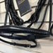 A black leather horse bridle with an attached bit and reins hanging on a retail display wall. A black hangtag with gold script branding is attached near the top.
