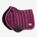 A quilted equestrian saddle pad in deep magenta-purple with white piping and a black leather LeMieux brand pad flap, shown on a light gray background.