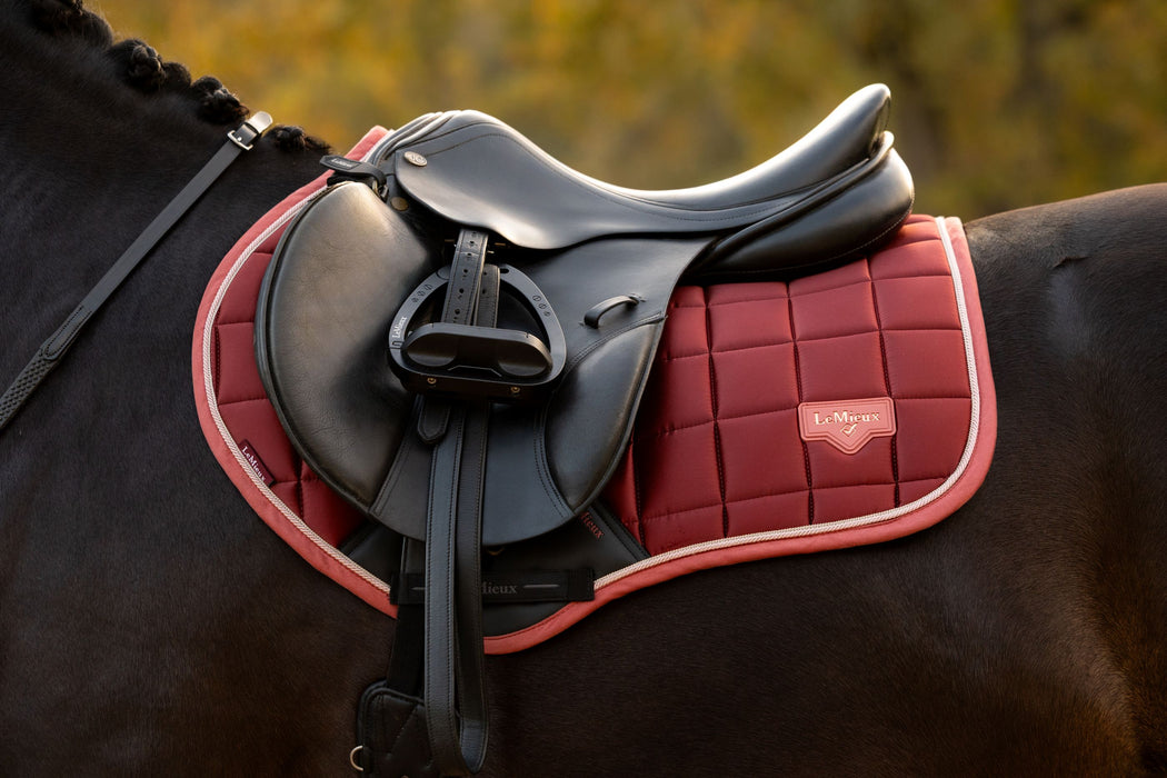 A close-up of a horse saddle resting on a orangish red quilted saddle pad with a black horse, set against a blurred outdoor background