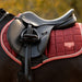A close-up of a horse saddle resting on a orangish red quilted saddle pad with a black horse, set against a blurred outdoor background