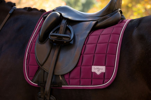A close-up of a horse saddle resting on a dark horse, showing a quilted magenta saddle pad with white piping and a small LaMoral logo patch.