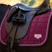 A close-up of a horse saddle resting on a dark horse, showing a quilted magenta saddle pad with white piping and a small LaMoral logo patch.