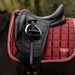 A close-up of an equestrian saddle with a red quilted saddle pad on a dark horse, featuring a small LeMieux logo patch on the pad.