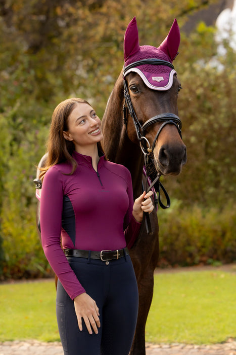 A woman in a purple long-sleeve riding shirt and navy breeches stands beside a brown horse wearing a purple fly veil and bridle, outdoors on a grassy field with trees in the background.