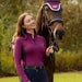 A woman in a purple long-sleeve riding shirt and navy breeches stands beside a brown horse wearing a purple fly veil and bridle, outdoors on a grassy field with trees in the background.