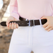 A close-up of a person wearing a dark brown leather belt with a silver-toned rectangular buckle, paired with white pants and a light pink long-sleeve top.