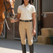 A woman stands in a stable holding a horse's reins, wearing a cream polo shirt, beige riding breeches with a black belt, and black riding boots.